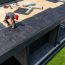 A construction worker is installing EPDM roofing material on a contemporary home under clear skies, surrounded by tools and prepared materials for the roofing project.